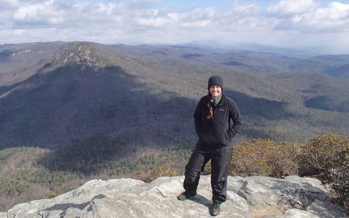 A person stands on a rock overlook and smiles at the camera. Behind them are the vast blue ridge mountains. 